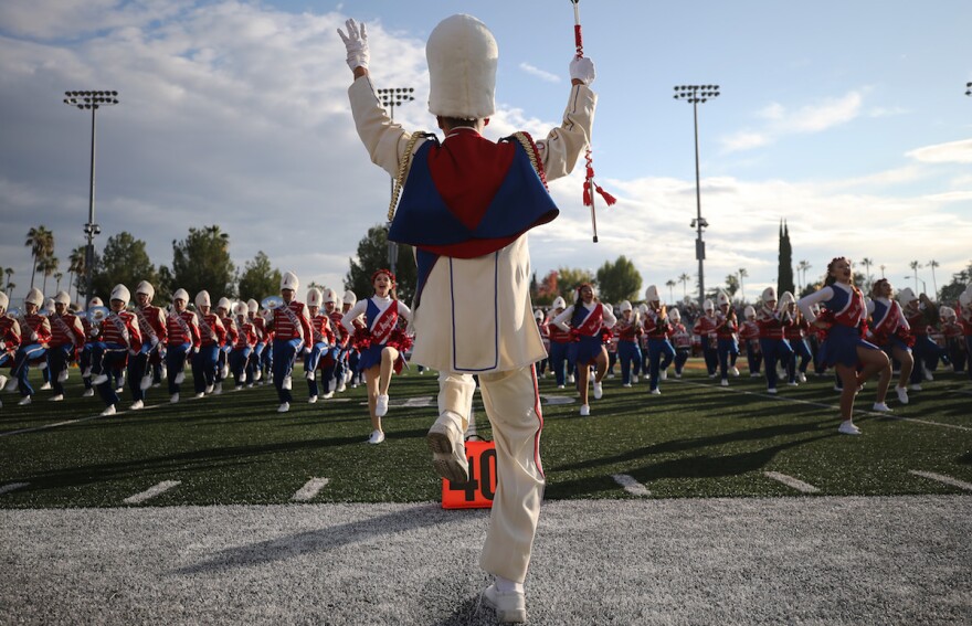 A high school marching band leader is shown from behind, with one foot up and their arms raised holding a baton, on a football field during the day. The leader is dressed in all white with a tall white hat and gloves, and a dark blue and red short cape. The band faces the leader in the background of the photo.