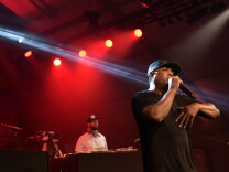 Rapper Chuck D, A middle-aged Black man, stands on a stage in front of a DJ and red lighting, holding a microphone. He wears a black t-shirt and a black hat. 