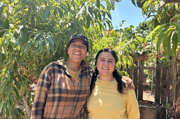 Two women are smiling at the camera with their arms around each other. The woman on the left is wearing an orange long-sleeve flannel and a gray baseball cap, while the woman on the right has her black hair pulled back in a ponytail and is wearing a pale-yellow shirt. They're both standing in an outdoor farm space filled with leaves and trees on a sunny, clear morning.