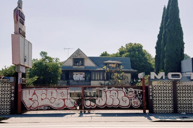 A wide view of the motel from the street. The gate between the breeze walls is closed. It's covered in heavy white and red graffiti. The dilapidated house is visible behind the gate, which is also covered in graffiti.