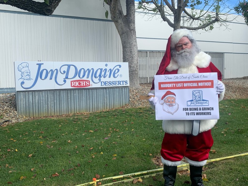 A man dressed as Santa Claus holds a sign saying "Naughty list official notice" in front of the Jon Donaire business sign.