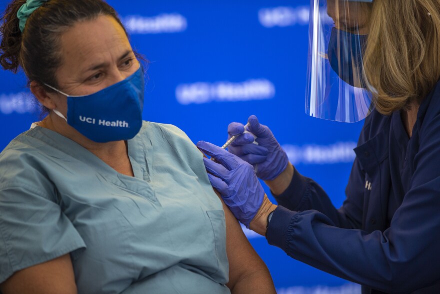 A woman wearing medical clothing and a face mask that says UC Irvine Health receives an injection in her left arm from another health care worker who is wearing rubber gloves, a face mask and face shield.