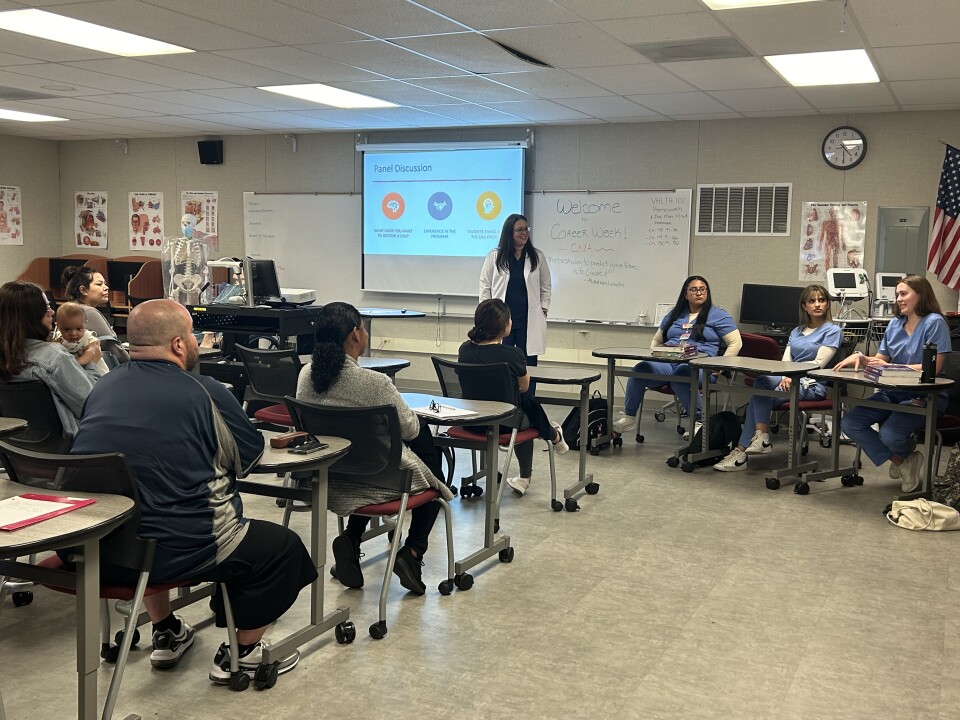 In a classroom decorated with posters about the human body, a handful of adults (and one baby) listen to three panelists and their professor. The panelists and the professor are wearing medical uniforms. 
