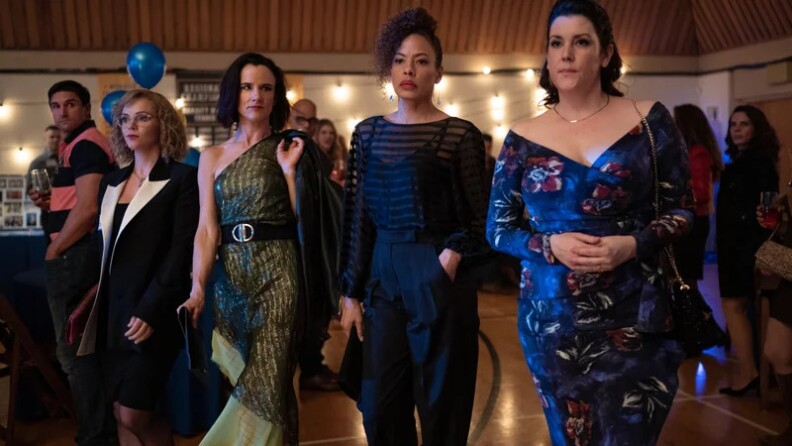 Four women who appear to be in their 40s or 50s enter an auditorium, dressed in cocktail attire. Three have light-tone skin and the fourth had medium-ton skin. They all look solemn.