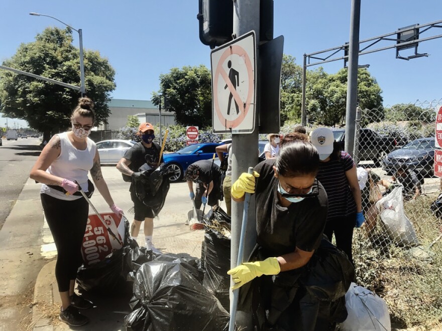 Volunteers gather their trash bags after combing the sidewalks along a Central Avenue intersection on a bright day. One worker has on a white tank-top and black pants, and the other has a black shirt. Both are wearing gloves.