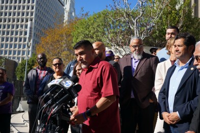 A man wearing red-collard shirt stands before a microphone stand. A crowd of people stand behind him. 