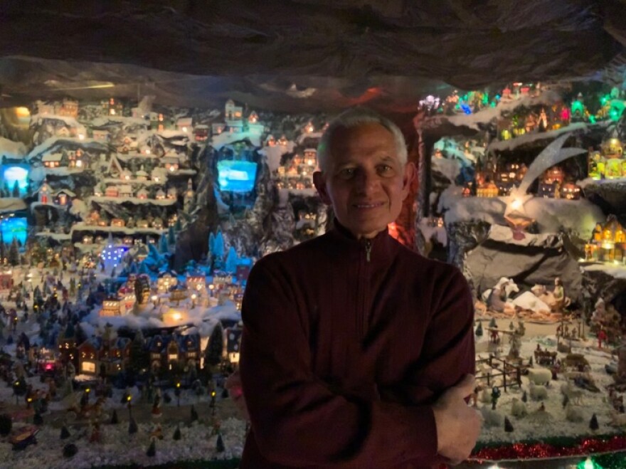 A man wit short white hair stands in front of a massive display of miniature Christmas houses.