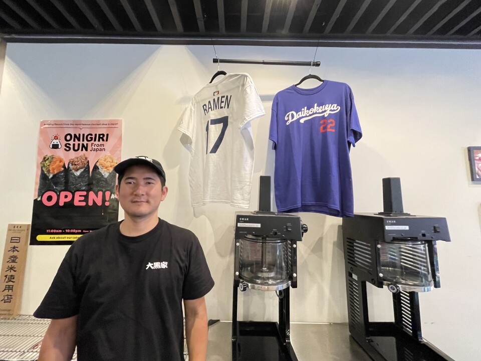 A Japanese man wearing a black t-shirt stands in front of two hanging t-shirts, one blue one that reads Daikokuya with the number 22, one white that reads 'ramen" with the No. 17.