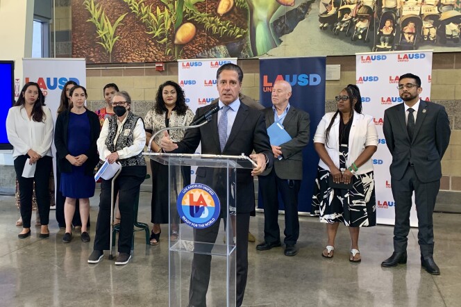 A group of people stand behind a man in a dark gray suit and a purple tie speaking at a lectern. Banners in the background read LAUSD. 