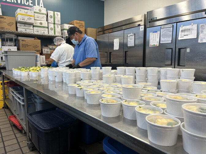 Two men in masks and hairnets are standing behind a metal kitchen counter. The counter is stacked with to-go containers filled with a mango salad.