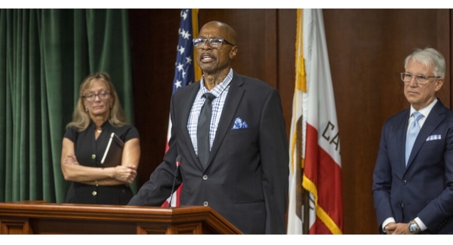 Maurice Hastings, a bald Black man with glasses wearing a dark suit and tie with a blue and white shirt and blue and white handkerchief in his breast pocket, speaks at a podium. Paula Mitchell, a white woman with her arms folded in front of her, stands to the left. DA George Gascón stands to the right.