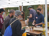 volunteers serve food at a table outside to unhoused people. There is a white tent covering the table.
