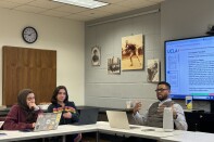 Three people sit in a small classroom, with their laptops in front of them. One of the people --- a man with medium skin tone, short dark hair, a long-sleeved shirt, and black-framed glasses-- gestures while explaining something.  Behind him, a document containing the words "Circumstances of Death" is projected on a large screen. 