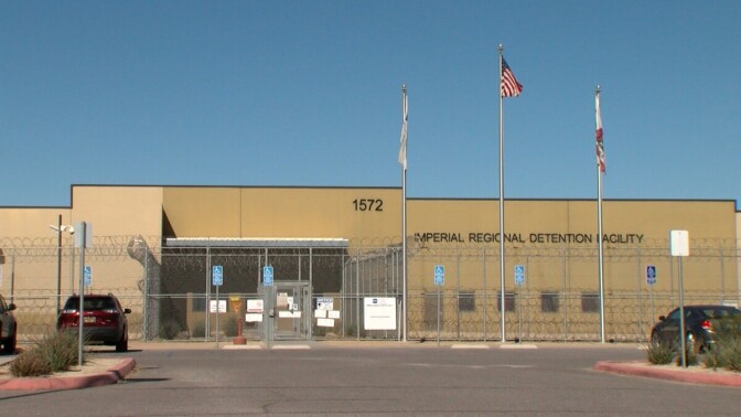 A one story, brown building behind a barbed wire fence. Three flagpoles are in front of the building. The middle flagpole is flying the American flag, the two white, unfurled flags hang on the other two poles. On the building is signage that reads, "Imperial Regional Detention Facility."