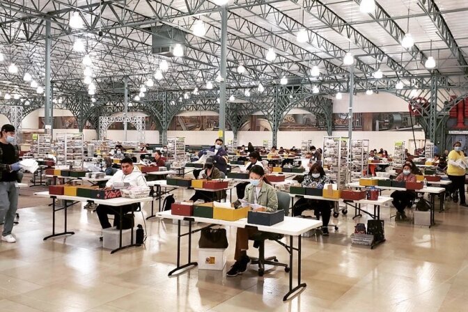 Election workers in a warehouse sit at voting tables and process votes