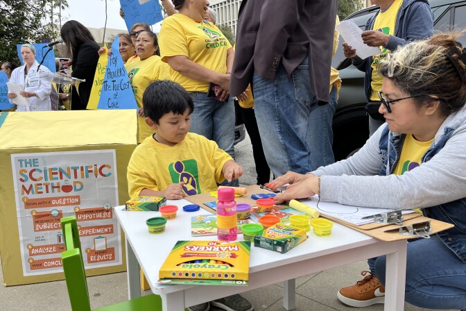 A 4-year-old boy with light brown skin plays with playdough at a children's table set up as part of a rally. People hold signs, chanting behind him. 