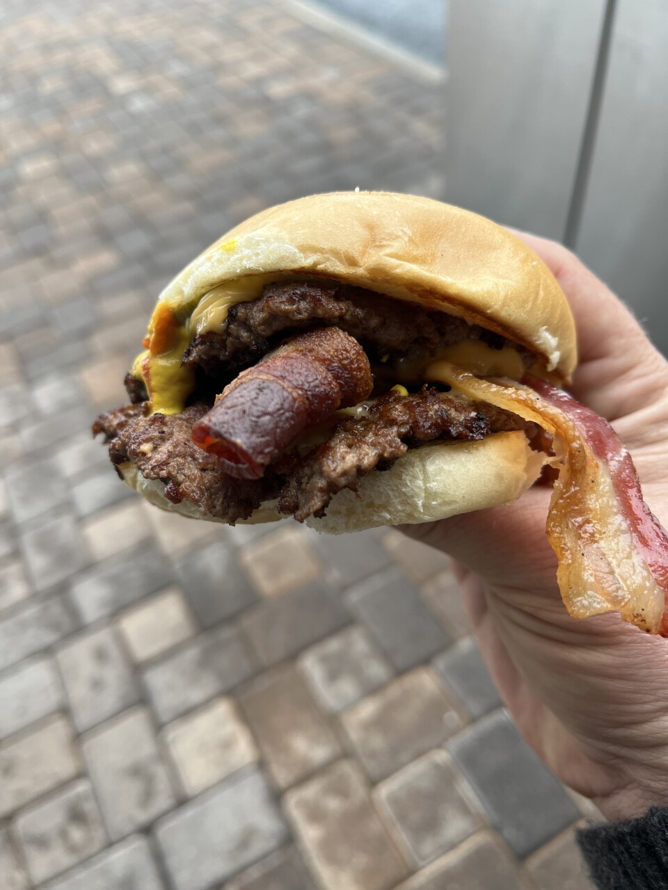 A hand with a light skin tone is holding a cheeseburger that features two large pieces of bacon protruding between the burger patties with a small amount of melted yellow American cheese.