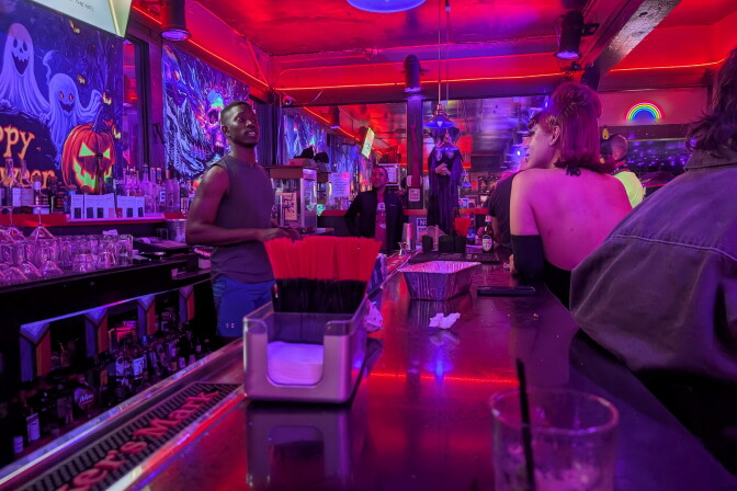 Interior of a bar with Halloween decorations. A black bartender is talking to a drag queen.