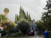 A rainbow arcs behind a small house and surrounding trees, the ground still wet with rain.
