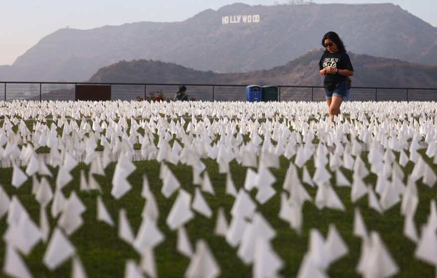 Scores of white flags are planted low on a grassy lawn with the Hollywood sign visible beyond in the hills. A person is walking through the flags.