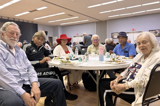 Six men and women, all older adults, are sitting around a white plastic table in a large interior room. White trays of food are placed in front of each of their seats.