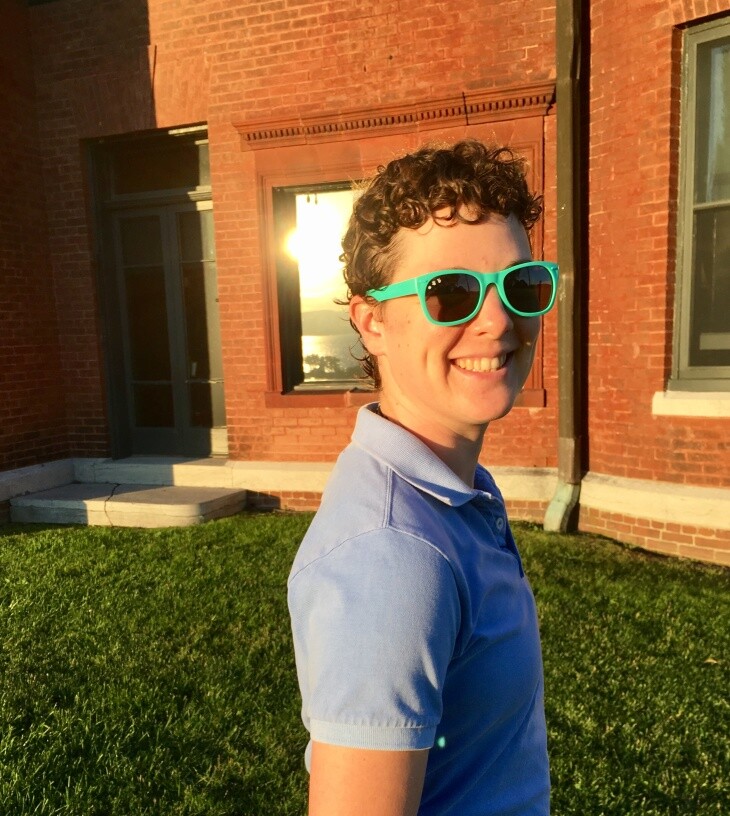 A person with light-tone skin, short hair and green-rimmed sunglasses smiles for the photo in a polo shirt