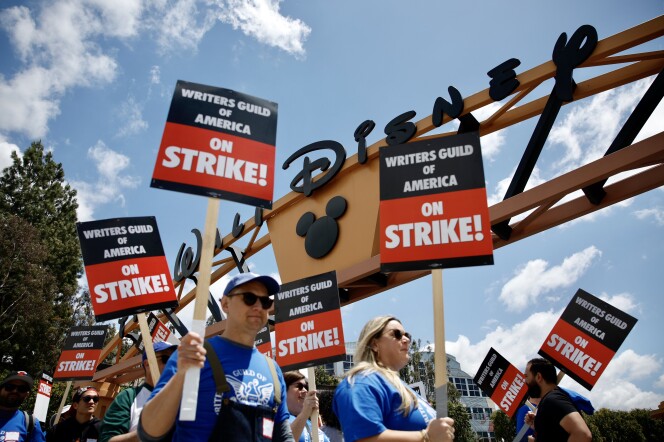 A group of people wearing blue T-shirts and holding black and white signs with the words "Writers Guild of America on Strike!" are picketing underneath a big sign that reads "Walt Disney." Scattered wisps of clouds break up the otherwise blue sky on a sunny day, and the man and woman closest to the viewer are wearing sunglasses. 