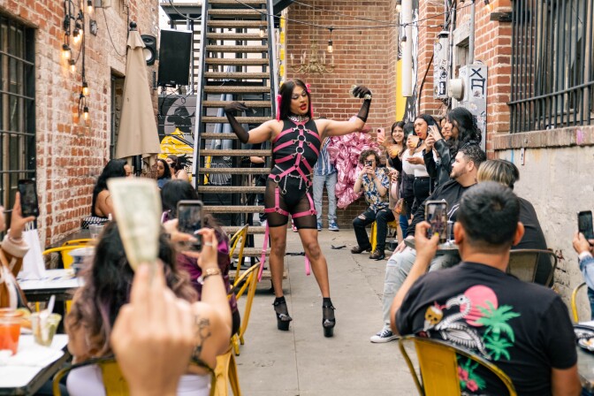 A drag queen wearing long black hair and pink and black outfit performs for a crowd in an outdoor patio. 