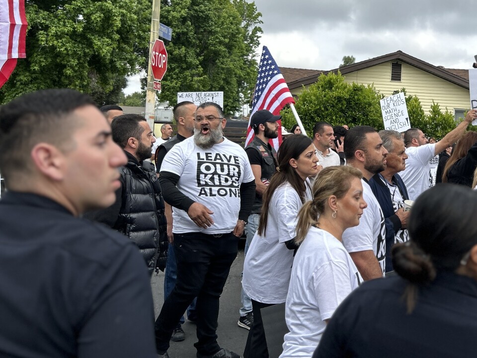A man with a medium-light skin tone stands in a crowd of protesters on the street wearing a black and white shirt that says leave our kids alone. In the foreground are police officers.
