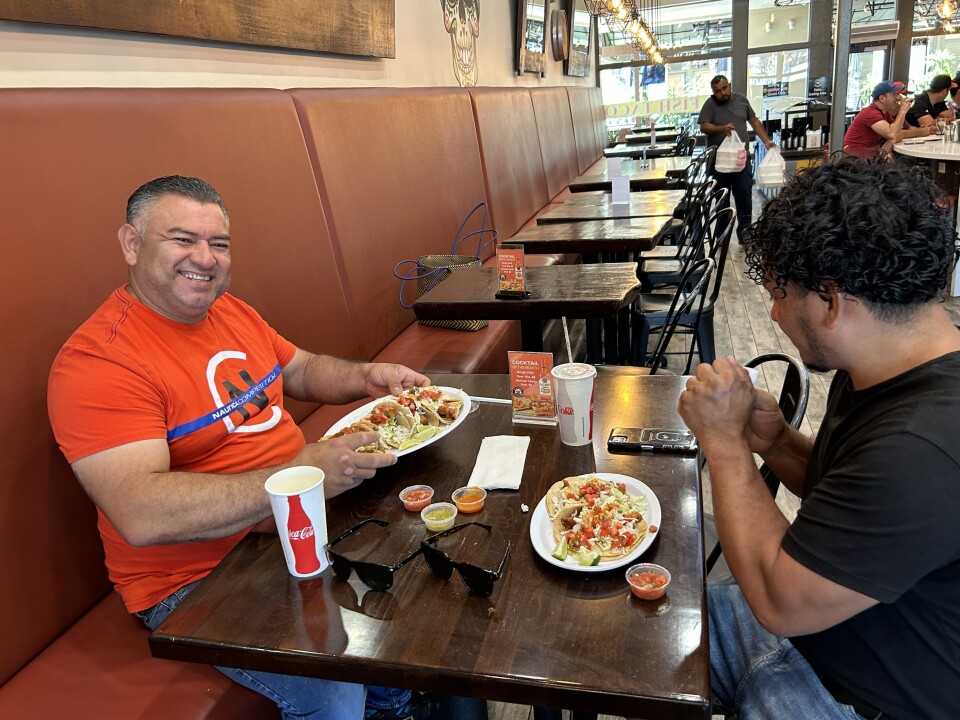 A man in an orange shirt shows off his plate of tacos and smiles for the camera. Another satisfied customer. He's sitting across the table with another man also enjoying tacos.