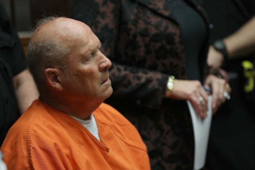 Joseph James DeAngelo sits in a orange jumpsuit and white undershirt. He looks forward, showing his right side profile. In the background is a women whose ring-adorned hands are clutching a piece of paper. Obscured behind her is a police officer.