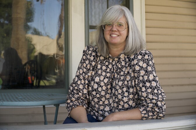 A light skinned woman with steel grey hair sits on a porch, facing the camera smiling. 