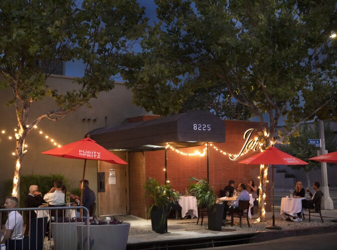 An image of the exterior of Jar, a restaurant located in the Beverly Grove neighborhood of Los Angeles. The outdoor dining area consists of various tables and chairs underneath red umbrellas, all positioned against the backdrop to the restaurant's front entrance, which is covered by a long black awing and red brick wall with the restaurant's illuminated signage. 
