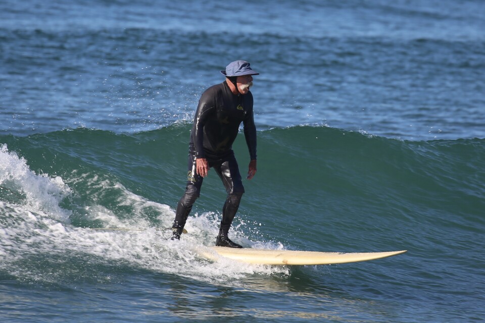 A man with a white mustache wearing a hat and wetsuit surfing a wave on a white board. 