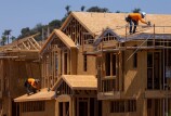 Two men wearing hard hats and long-sleeved bright orange shirts bend over to work on the wood frame roof of a housing complex under construction.