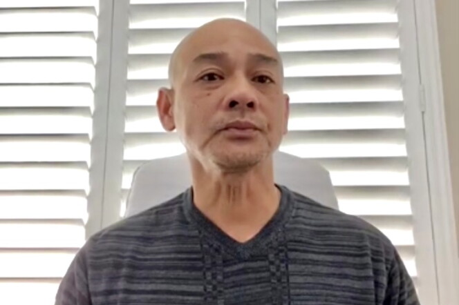 A 48-year-old man of Vietnamese descent with a shaven head looks somberly into the camera, with a window with slatted blinds behind him. 