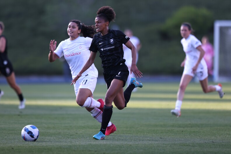 A soccer play dribbles the ball down the field while a defender chases after them. Other players run behind them in the distance.