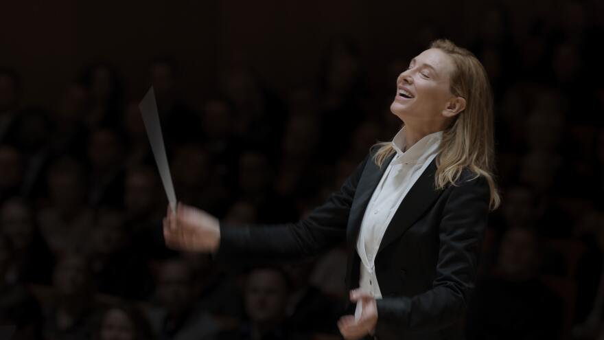Kate Blanchett stands right of center. Her eyes are softly closed as she smiles with her head slightly raised upwards giving an air of euphoria in her expression. She wears a tailored black suite with a white waistcoat and shirt. Blanchett's left arm is raised up by her waist while her right arm holds a baton slightly higher. Her arm and baton are blurred from the quick motion of her waving it. Blanchett stands in front of a dark background, but hazy outlines of audience's faces can be seen behind her.