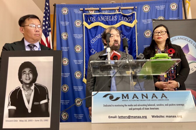 A Japanese American man speaks at a podium, flanked by a Chinese American man and a Chinese American woman. A black and white photo of an Asian American man is placed at the forefront. 