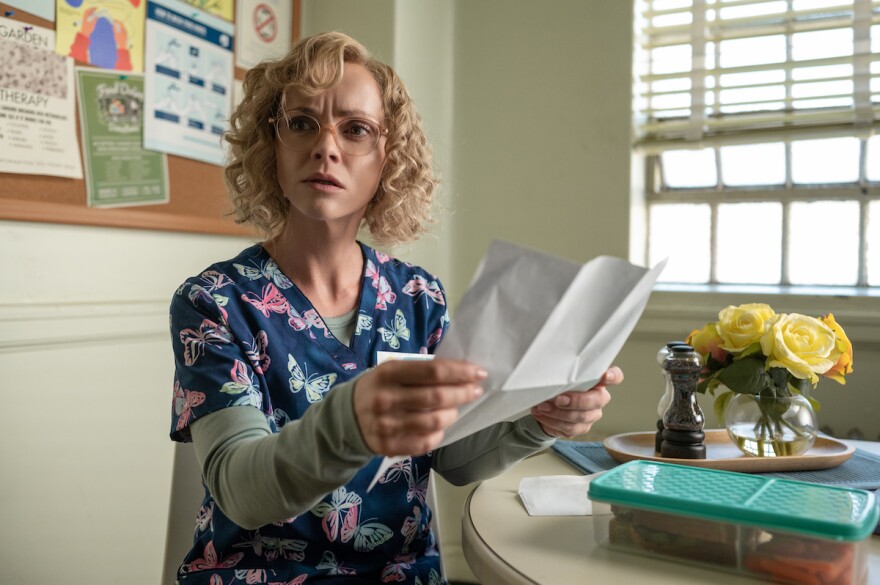 Christina Ricci sits at a table, wearing floral scrubs, holding a paper in her hand.