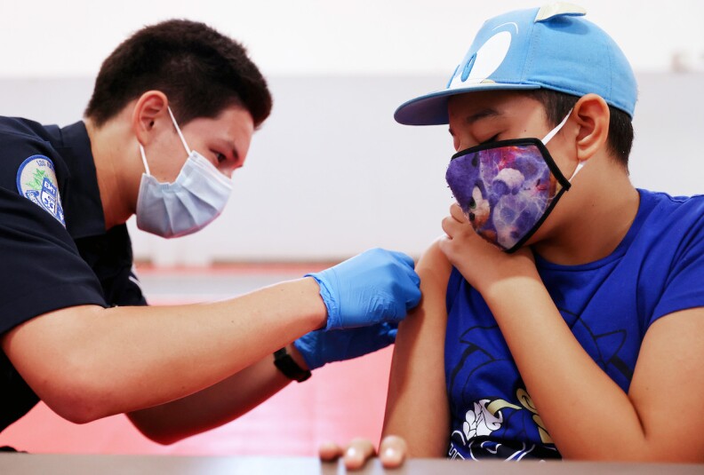 a man in a mask an dark uniform puts a band-aid on the arm of a young man, also in a mask