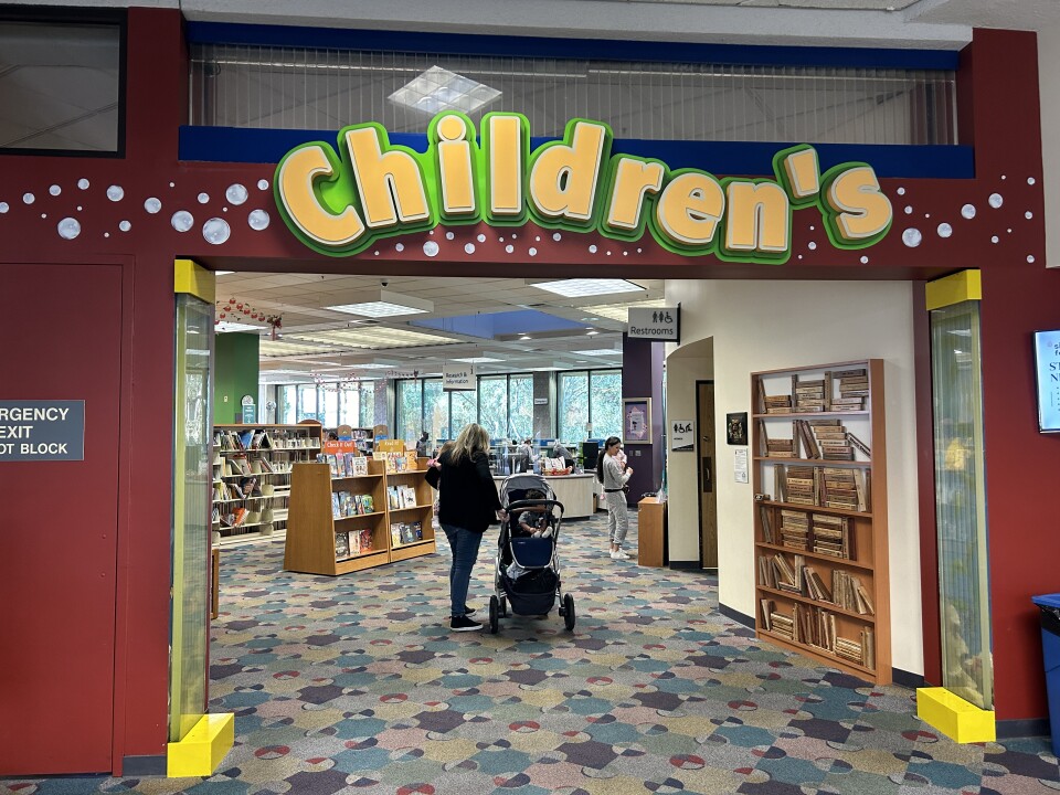 An entryway with a large yellow sign above that reads "Children's." Through the entry, there's a female-presenting person next to a baby stroller and shelves of books in the distance. 