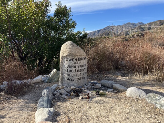 A gray headstone sits on gravelly soil, in front of dry brush and a tree. The headstone reads "Owen Brown, son of John Brown The Liberator, Died Jan. 9, 1889, aged 64 yrs." Smaller stones and pine cones are gathered at the base of the headstone. Mid-sized gray stones mark the ground in front of the headstone into the shape of a grave. The sky is blue and there are hills and telephone lines behind the scene.