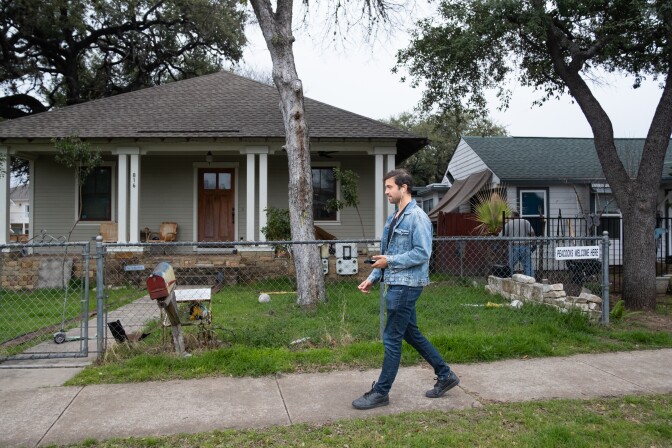 A man with light-ton skin walks past a home with a chainlink fence and a front porch.