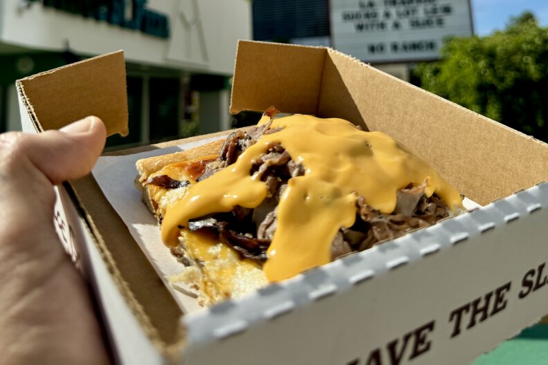 A small pizza box with a square slice of pizza with beef and melted cheese on top is being held up by a hand outside of a storefront and a facing an Arby's Roast Beef hat-shaped sign.