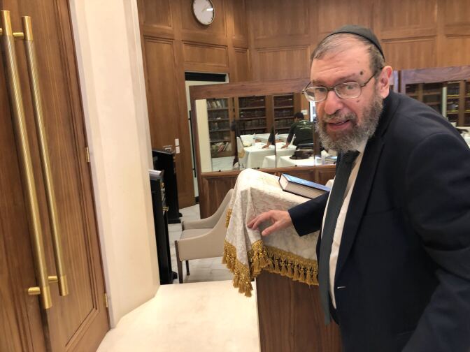 A man with light-tone skin has a full beard and glasses. He wears a dark suit and tie with a skull cap. Behind him is a book on a lectern covered with a tassled cloth and more book shelves are visible in the background.
