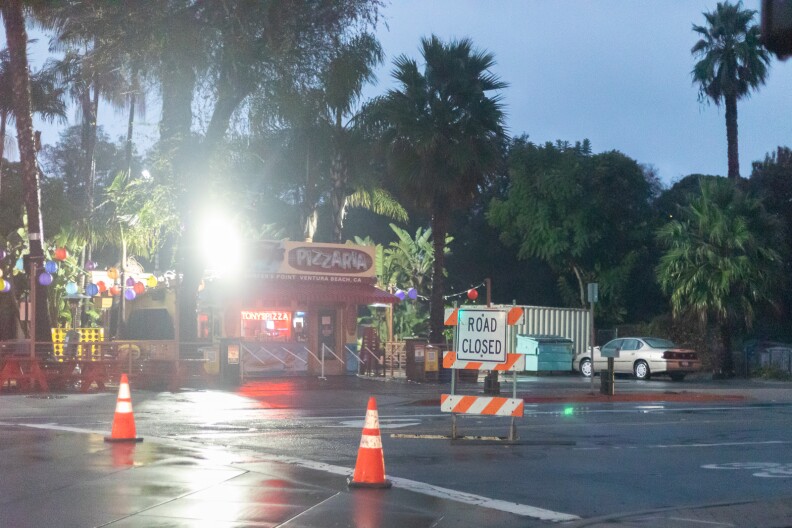 A structure with a red sign that says "Pizzaria." On the wet road in front of the pizza place there are two orange cones and a sign that says "road closed." Trees in the background and palm trees next to the pizza shop.