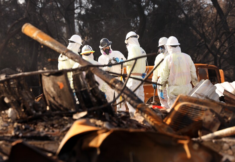 People in protective suits and other safety gear stand around a burned down home.