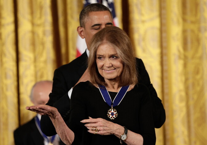 A man wearing a black suit stands behind a woman wearing a black dress. The man is placing a medal hanging on a blue ribbon around her neck. Behind them is a gold curtain.