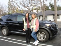 The mental health crisis response workers stand in front of a black SUV. The wear reflective safety vests and are looking towards the camera. 